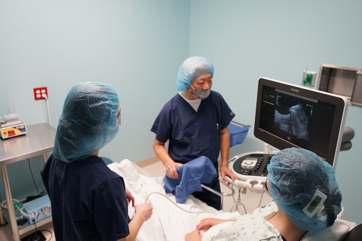 A woman is getting an ultrasound from two medical professionals standing around her bed.